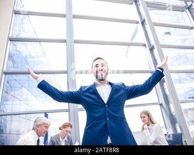 Junge freudige Geschäftsmann stehend mit erhobenen Armen lächelnd, Team von seinen Kollegen an dem Tisch im Hintergrund sitzen Stockfoto