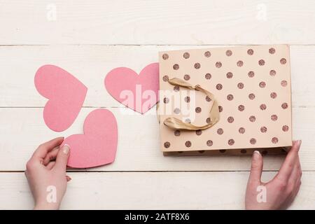 Weibliche Hand mit pinkfarbenem Herzen. Papiertasche mit Valentinsgruben im Vintage-Hintergrund. Liebe und Urlaubskonzept. Valentinstag Karten oder Dekorationen, Draufsicht. Stockfoto