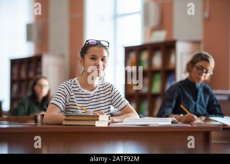 Lächelnde Teenager in der Casualwear, die Sie beim Sitzen in der Bibliothek betrachtet Stockfoto