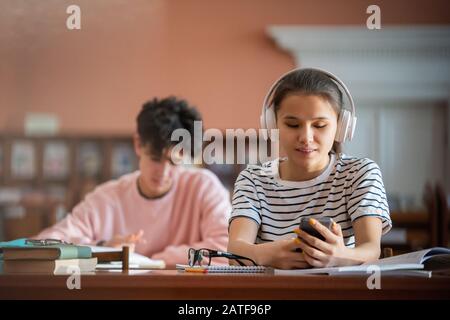 Hübscher Student mit Kopfhörern scrollt in Smartphone in Bibliothek Stockfoto