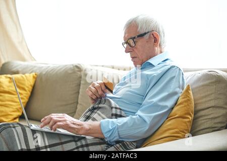 Der pensionierte grauhaarige Mann ist in eine entspannte Atmosphäre auf der Couch eingewickelt und online einkaufen Stockfoto