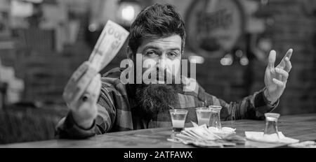 Hiphster hält Geld, Bargeld, um mehr Alkohol zu kaufen. Mann mit betrunkenem Gesicht sitzt allein an der Theke. Betrunkenes Konzept. Guy verbringen Sie Freizeit in der Bar mit einem Cocktail, unverschäumter Hintergrundfarbe. Stockfoto