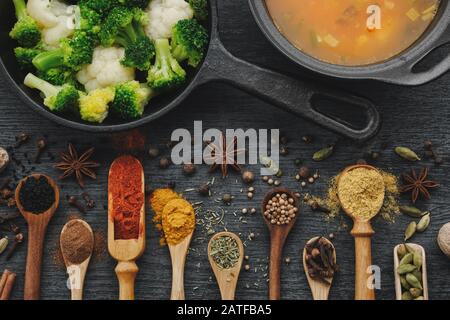 Verschiedene aromatische, bunte Gewürze und Kräuter in Holzlöffeln und Schaufeln. Brokkoli und Blumenkohl auf eiserner Bratpfanne. Schwarzer Keramiktopf mit Suppe Stockfoto