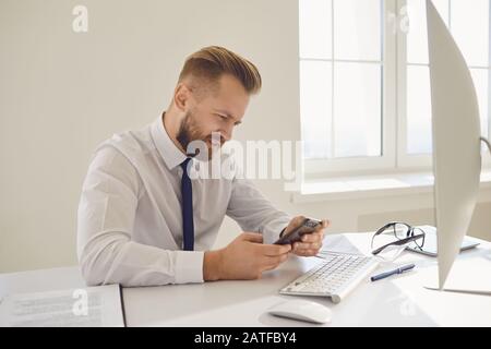 Ein Unternehmer, der mit viel beschäftigt ist, liest die Nachricht am Telefon an einem Tisch mit einem Computer im Büro. Stockfoto