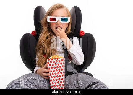 Ein kleines Mädchen mit Makeup und langen blonden Haaren sitzt auf einem Autokabystuhl und sieht Film oder Cartoon mit Popcorn isoliert auf weißem Hintergrund aus. Stockfoto