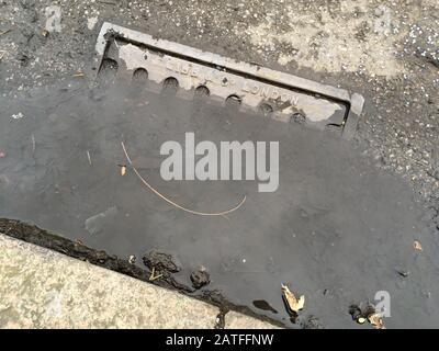 Wasserpfütze um eine alte Eisenstraße / Straßenabflussgitterabdeckung, bei der die fehlenden Wartungs- und Räumungsarbeiten die Abflüsse blockiert und das Regenwasser nicht mehr mitführen können. GROSSBRITANNIEN. Stockfoto