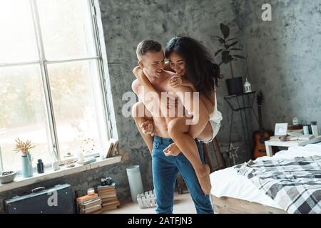 Gemischtes Rennpaar. Junger Mann, der Frau wieder im Schlafzimmer trägt und freudig lachte Stockfoto