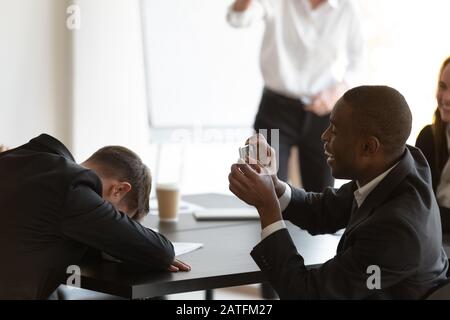 Mitarbeiter, der während der Briefing schläft, afrikanischer Kollege fotografiert von ihm Stockfoto
