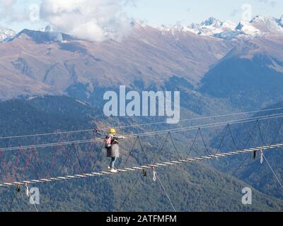 Ein Mädchen in einem gelben Helm und mit Rucksack überquert eine Hängebrücke über eine Klippe auf dem Grund verschneiten Berggipfels Stockfoto
