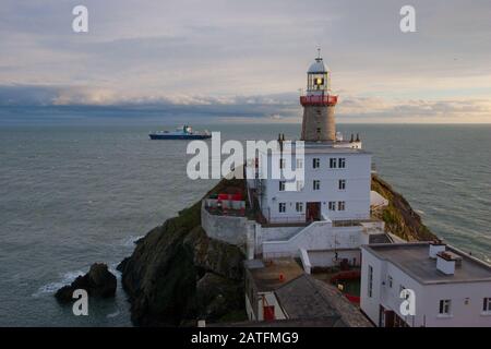 Der Baily Leuchtturm, Howth. co. Dublin, Baily Leuchtturm auf Howth Klippen, Blick auf den Baily Leuchtturm von der Klippe mit Frachtschiff im Hintergrund Stockfoto