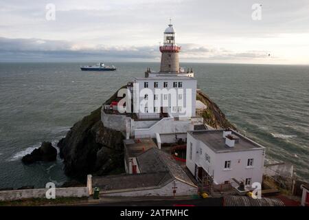 Der Baily Leuchtturm, Howth. co. Dublin, Baily Leuchtturm auf Howth Klippen, Blick auf den Baily Leuchtturm von der Klippe mit Frachtschiff im Hintergrund Stockfoto