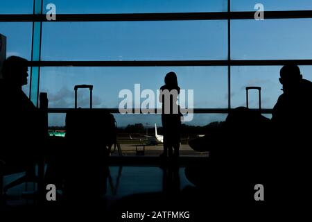 Silhouette von Fluggästen in einer Flughafenlounge am breiten Beobachtungsfenster, die ein Flugzeug vor einem surrealen Sonnenuntergang fliegen sehen. Dublin Stockfoto