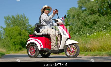 Portrait einer afroamerikanischen Frau in Sonnenbrille und einem elektrischen Roller mit Hut. Freiheit des Fahrens von Mobility Vehicle entlang des grünen Stadtparks auf Summ Stockfoto