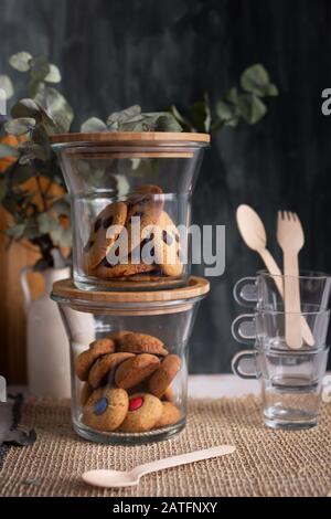 Nahaufnahme eines Haufens von Glasgefäßen voller hausgemachter Schokoladenchip-Plätzchen. Eine Holzgabel und ein Löffel befinden sich in einem anderen Stapel Kaffeetassen Stockfoto