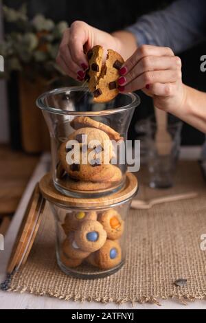 Nahaufnahme eines Haufens von Glasgefäßen voller hausgemachter Schokoladenchip-Plätzchen. Ein Plätzchen wird von den Händen der Frau gehalten. Stockfoto