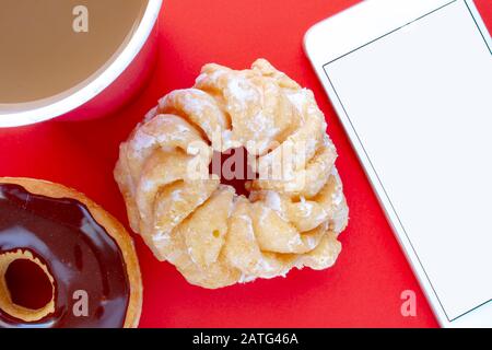 Eine Tasse Kaffee mit einem Chocolate Dip Donut und Honey Cruller Donut mit einem Smartphone Stockfoto