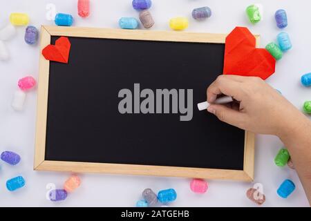 Die weibliche Hand hält Kreideschrift auf Tafel mit roter Herzform und buntem Glitter über dem weißen Hintergrund. Grußkarten, Liebe und Vale Stockfoto