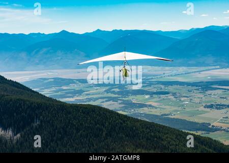 Der Mann fliegt mit seinem Hang an klarem sonnigen Tag über ländliche Felder. Langer Schuss. Schöne Kootenay-Tal-Berge im Hintergrund Stockfoto