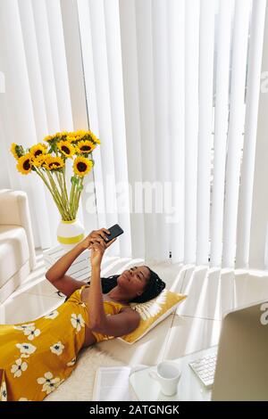Liebenswürdig lächelnde junge Frau, die zu Hause auf dem Boden liegt und Textnachrichten liest, bilden ihren Freund Stockfoto