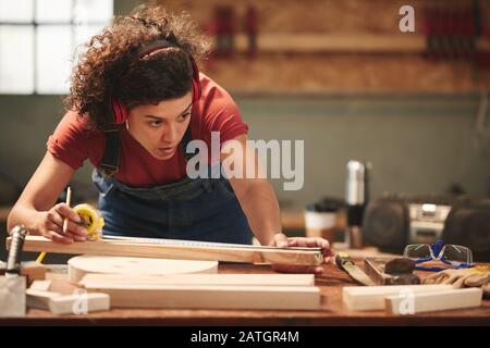 Junge konzentrierte geschweifte Frau in Ohrenschützern und insgesamt messende Holzplanke mit Maßband Stockfoto