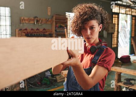 Junge konzentrierte Frau mit lockigen Haaren, die Holzpflaster hält und ihre Länge mit den Augen misst Stockfoto