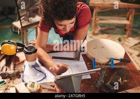 Hochwinkelige Ansicht der jungen Tischlerinnen, die das Handbuch auf der digitalen Tablette lesen, während sie sich über einen Tisch lehnen, der mit Zugluft, Werkzeug und Sägespänen bedeckt ist Stockfoto