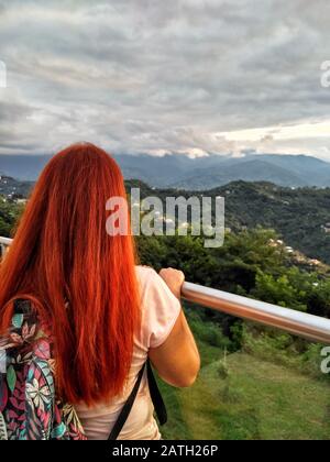 Der Rücken eines rothaarigen Mädchens steht auf einer Aussichtsplattform und blickt auf die Berge und die erstaunliche Natur. Reise und Urlaub, vertikales Foto Stockfoto