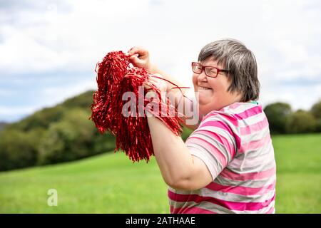 Geistig behinderte Frau jubelt mit roten Pompons auf einer grünen Wiese und hat viel Spaß Stockfoto