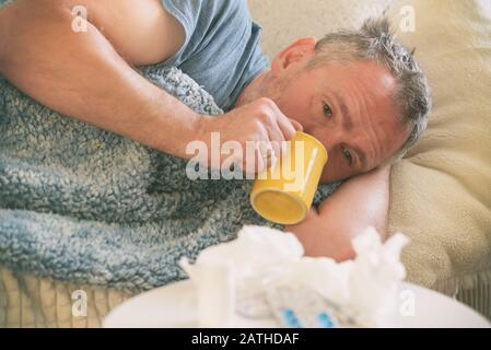 Der an Coronavirus, Grippe oder Kälte leidende Mann liegt leider mit einer Decke auf seinem Bett bedeckt. Medikamente, Medikamente, Becher und Gewebe im Vordergrund Stockfoto