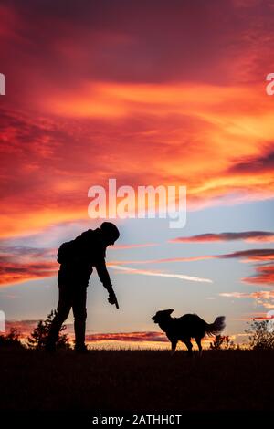 Silhouetten eines Mannes und seines Hundes vor Sonnenuntergang oder Sonnenaufgang, rote Wolken Stockfoto