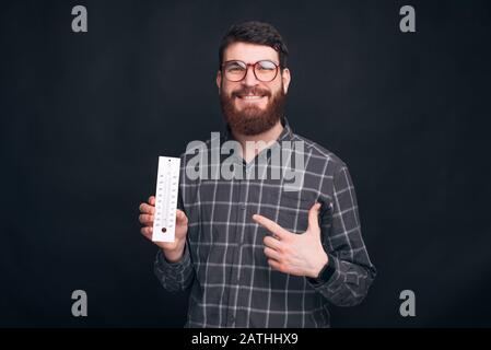 Der junge Mann, der eine Brille trägt, hält ein Thermometer und zeigt darauf. Stockfoto