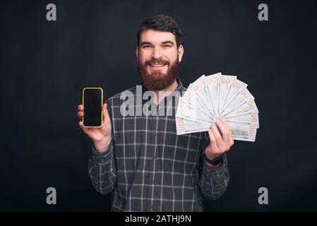 Der junge bärtige Mann hält sein Telefon und einen Haufen Geld in der anderen Hand. Sie können über Ihr Telefon arbeiten. Stockfoto