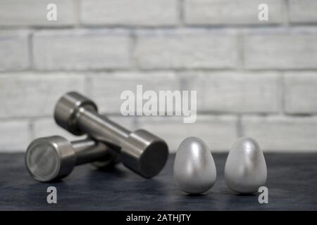 Metall-Hanteln und Stahleier gegen eine weiße Ziegelwand. Das Konzept von Beharrlichkeit und Effizienz über sich selbst. Stockfoto