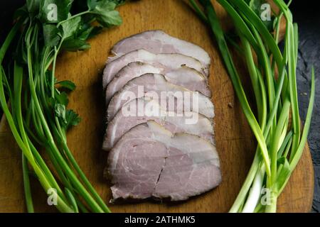 Geschnittener Schinken auf einem Holzbrett, um den Schinken herum, grüne Zwiebeln, Dill und Petersilie. Stockfoto
