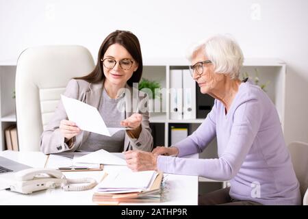 Lächelnde junge Sozialarbeiterin, die mit der Seniorin am Tisch sitzt und ihr Dokumentinhalt erklärt Stockfoto