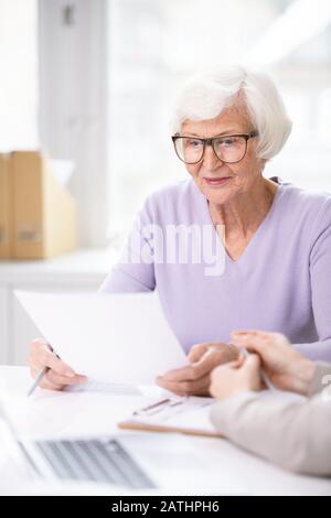 Konzentrierte ältere Frau in einer Brille, die am Tisch sitzt und vor der Unterzeichnung das Dokument liest Stockfoto