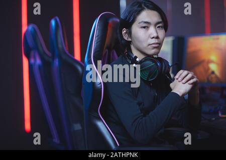 Portrait des jungen Asiaten, der auf die Kamera schaut, während er im Gaming-Stuhl im dunklen Cyber-Sport-Innenraum sitzt, kopiere Platz Stockfoto