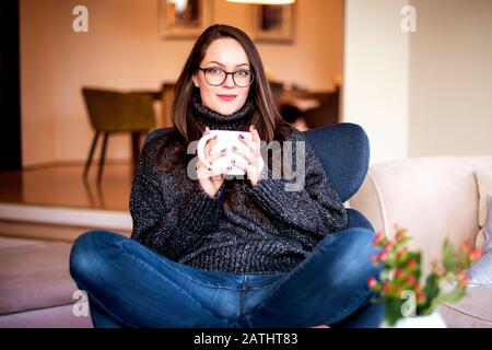 Portrait einer attraktiven jungen Frau, die ihren Tee trinkt, während sie sich zuhause im Sessel entspannen kann. Stockfoto