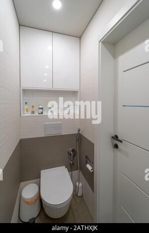 moderne designtoilette mit beigen und braunen Wand- und Bodenfliesen, Holztüren, weißer integrierter toilettenschüssel, bin, bidet Dusche, Schrubbbbürste und Lampe in Stockfoto