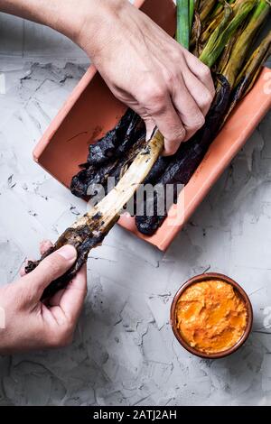 Mann schält ein gegrilltes calcot, eine für Katalonien typische süße Zwiebel, Spanien, neben einer Steingutschüssel mit Romesco-Sauce, um darin zu tauchen, auf einem gruseligen Rusti Stockfoto