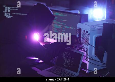 Seitenansicht bei nicht erkennbarem Cyber-Security-Hacker, der Kapuze trägt, während er an einem Computersystem in dunklem Raum arbeitet, Kopierraum Stockfoto