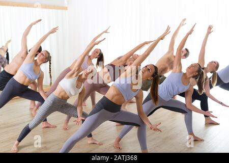 Gruppe von jungen sportlich attraktive Leute im Yoga Studio, Yoga Unterricht mit Kursleiter, Stretching auf Fußboden im Trikonosana, Dreieck yoga Pose Stockfoto