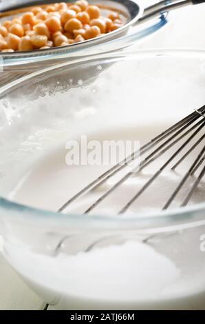 Glasküssel aus gepeitschtem Aquafaba (veganer Eiweiß-Ersetzer) im Vordergrund mit Ballonwisk. Kichererbsen (Garbanzo-Bohnen), die durch Metallsieb strapazen Stockfoto