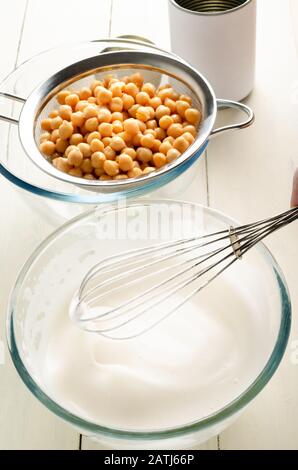 Abgewinkelter Schuss aus gepeitschtem Aquafaba in Glasschale mit Schneebesen und abtropfenden Kichererbsen im Sieb. Eine leere, nicht gebrannte Zinn sitzt im Hintergrund auf weiß gestrichenem Stockfoto