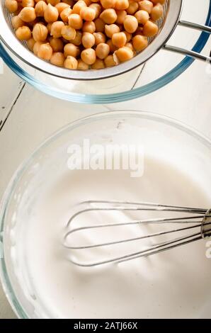 Overhead-Aufnahme von gepeitschtem Aquafaba in einer Glasschale mit Ballonwisk und Kichererbsen, die durch das Metallsieb auf einem bemalten weißen Holztisch gehobelt werden. Stockfoto