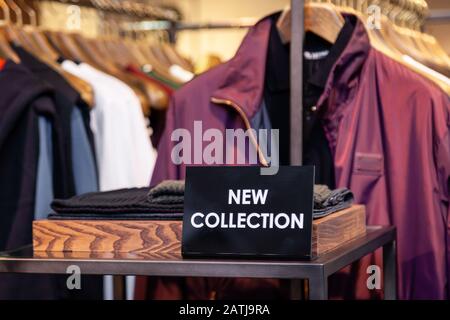 Platte mit Aufschrift neue Kollektion auf Boutique-Regalen, Kleiderständer mit bunten männlichen, weiblichen Kleidungsstücken auf Bügeln. Konzept Luxus-Shop, Shopping Stockfoto