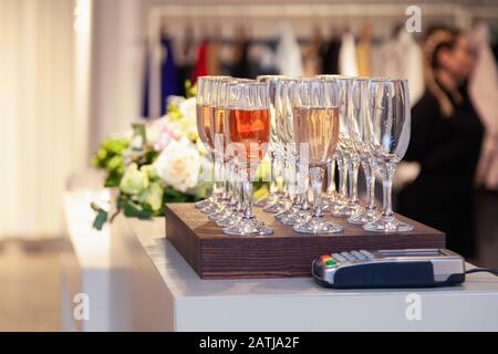 Blumen und Weingläser mit Rose und weißem Champagner stehen in Reihe auf Holzschreibtisch auf der Präsentation des Bekleidungshauses Stockfoto