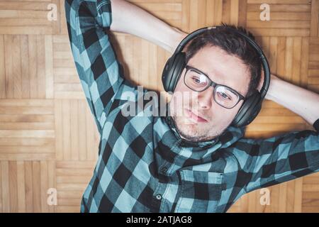 Der junge kaukasische Mann mit kariertem blauem Hemd liegt auf dem Holzboden und zählt Musik mit kabellosen Kopfhörern auf Stockfoto