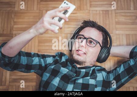 Der junge kaukasische Mann mit kariertem blauem Hemd liegt auf dem Holzboden und zählt Musik mit kabellosen Kopfhörern auf Stockfoto