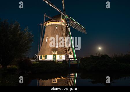 Die traditionelle holländerwindmühle wird nachts beleuchtet. Steigender Vollmond im Hintergrund. Stockfoto